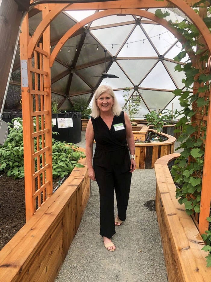 IMG_20190806_113157_528 - Growing Spaces Greenhouses A smiling woman standing in a greenhouse, with lush raised garden beds on either side, framed by a wooden arch with a geodesic dome ceiling in the background.