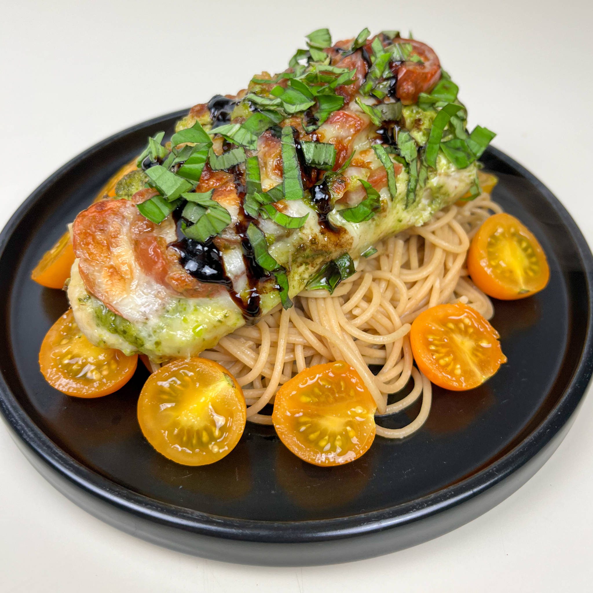 Pesto Caprese Chicken Garnished with Fresh Basil, Cherry Tomatoes, and Balsamic Glaze Served Over Buttered Whole Grain Spaghetti Noodles