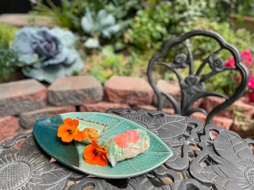 dome_grown_recipe_nasturtium_spring_rolls - Growing Spaces Greenhouses nasturtium spring roll on a green leaf-shaped plate, garnished with bright orange nasturtium flowers on a wrought iron table in a Growing Dome