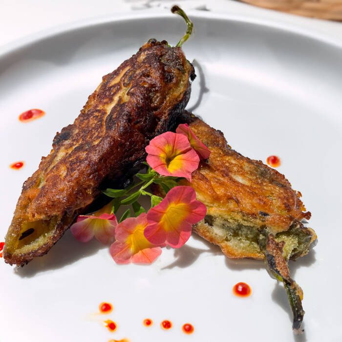 fried green chile rellenos on a white plate garnished with peach-colored petunias and red Chile drizzle