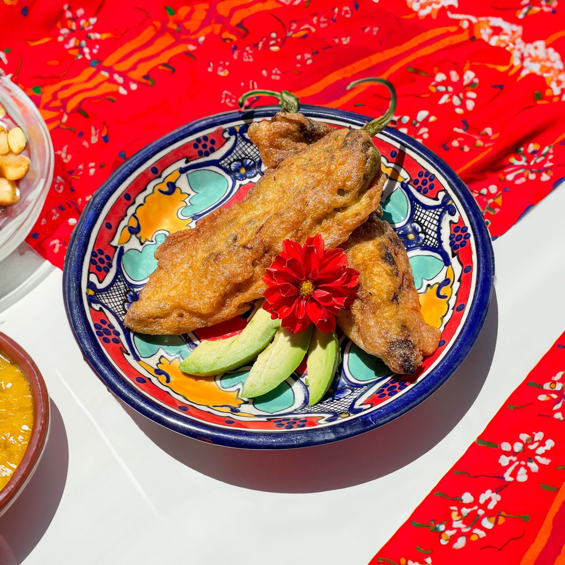 wild_rice_mushroom_stuffed_green_chile_rellenos - Growing Spaces Greenhouses Green Chile Rellenos garnished with avocado slices and a red dahlia on a talavera plate set on a red table cloth