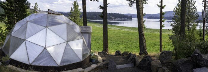 panoramic view of the exterior of a 22' Growing Dome with open upper vents, surrounded by tall evergreens, green marsh, and lake in background