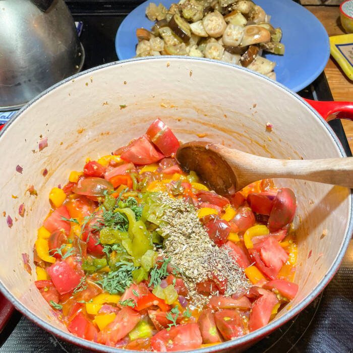 a pot on the stove full of tomatoes, herbs and chiles with a plate of cooked eggplant in the background