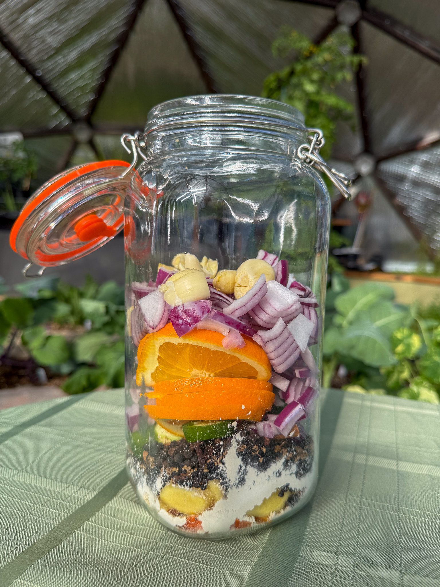 A jar filled with fire cider recipe ingredients waiting to be filled with apple cider vinegar in a geodesic greenhouse